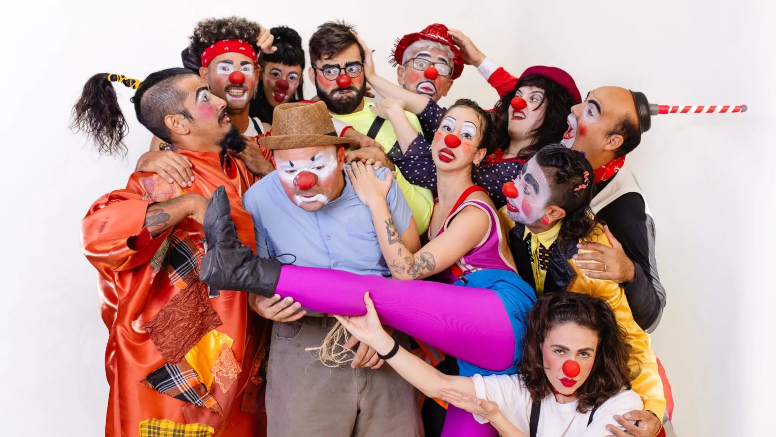 Fotografia em grupo de dez pessoas caracterizadas como palhaços, com narizes vermelhos e maquiagens faciais expressivas. Elas posam juntas, sobrepostas, olhando para a câmera contra um fundo branco.